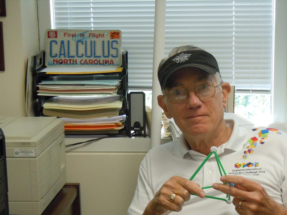 Harold Reiter in his office holding a mathematical model with a NC license plate behind him that says "calculus"