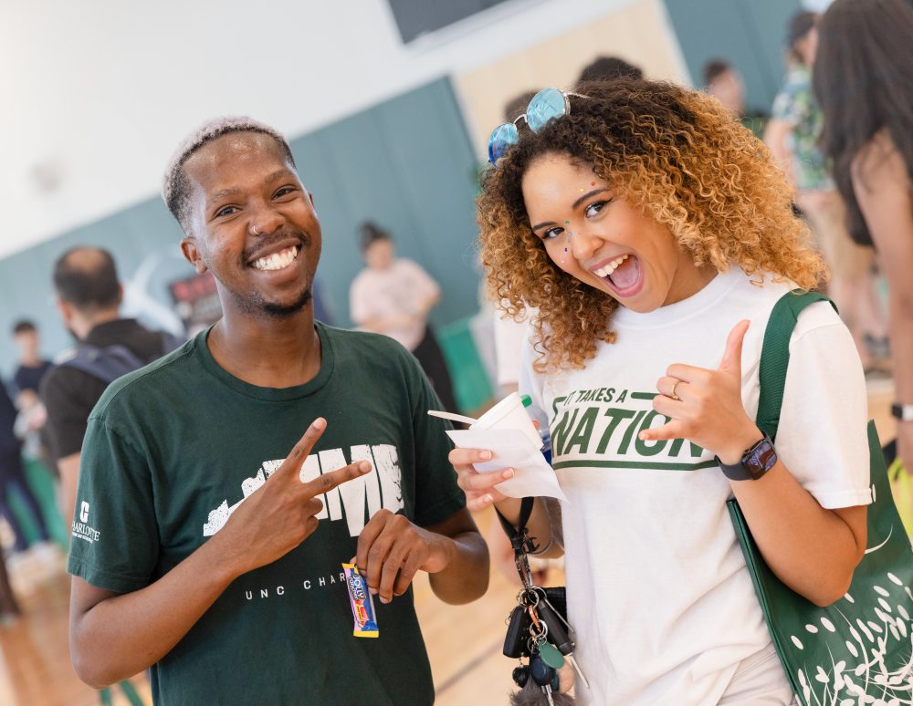 Two students in Niner gear smile and hold up pickaxe and peace hand signs.