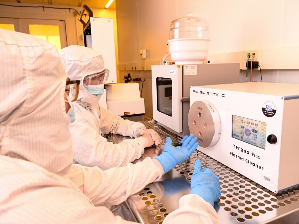 Two people in full bosy clean room suits and gloves work in front of a plasma cleaner while holding a lens.