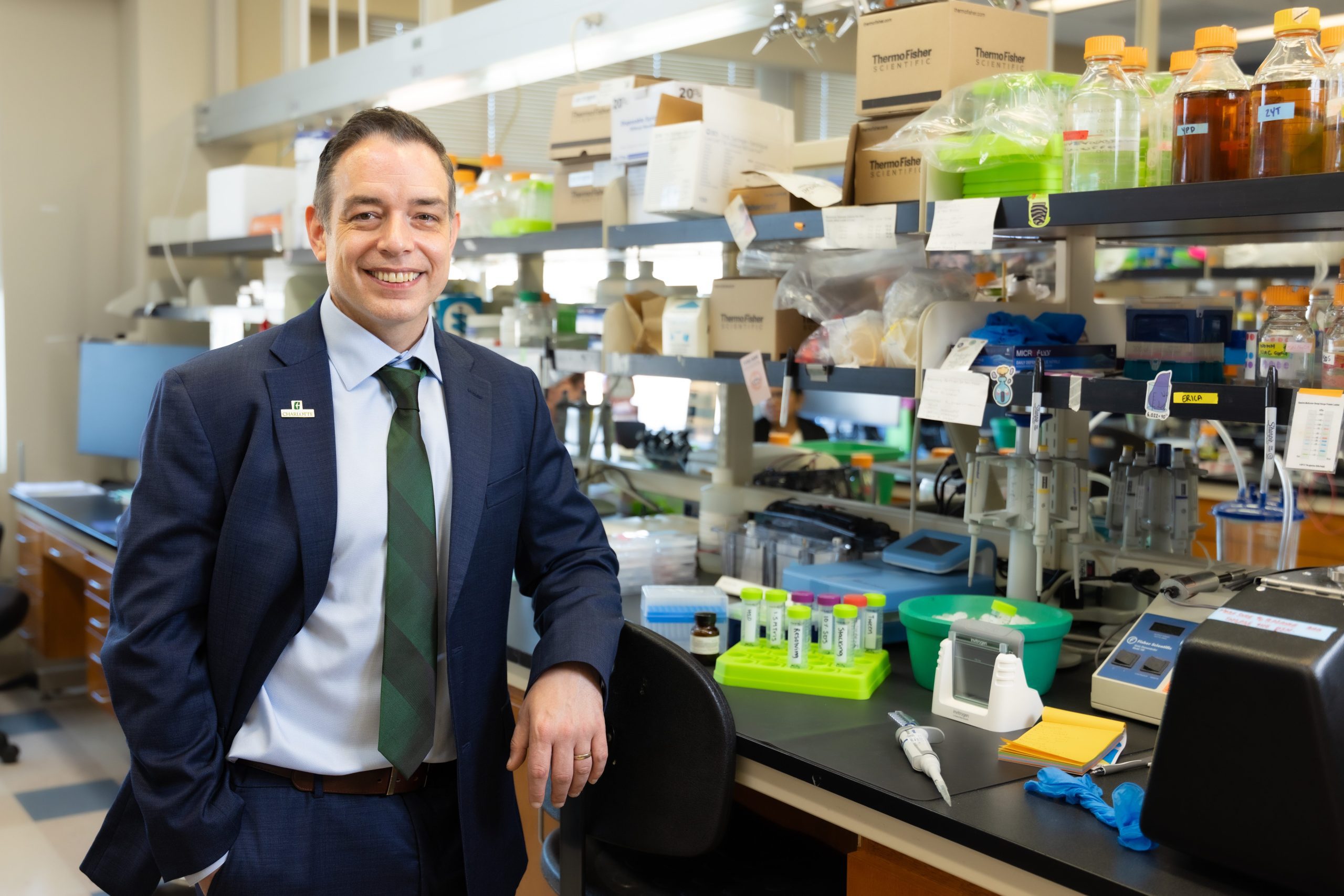 Andrew Truman stands in his biology lab, wearing a navy suit, green tie, and a UNC Charlotte pin. He is leaning against a lab chair and smiling at the camera.