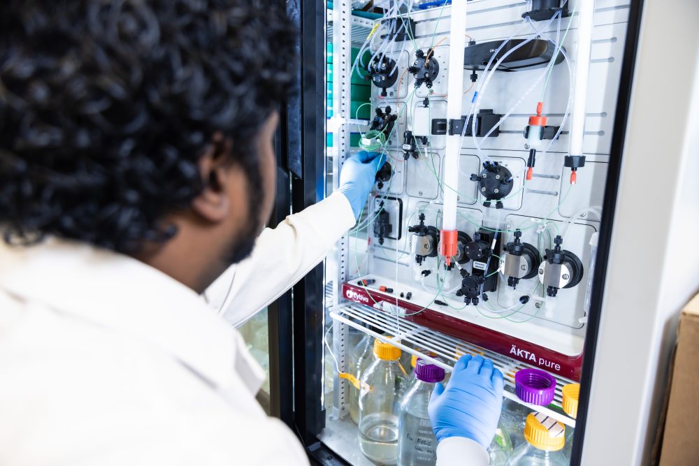 In the foreground, the back of a researcher's head as he reaches into a cooler containing an ӒKTA pure™ chromatography system. The researcher is wearing a white lab coat and blue gloves.