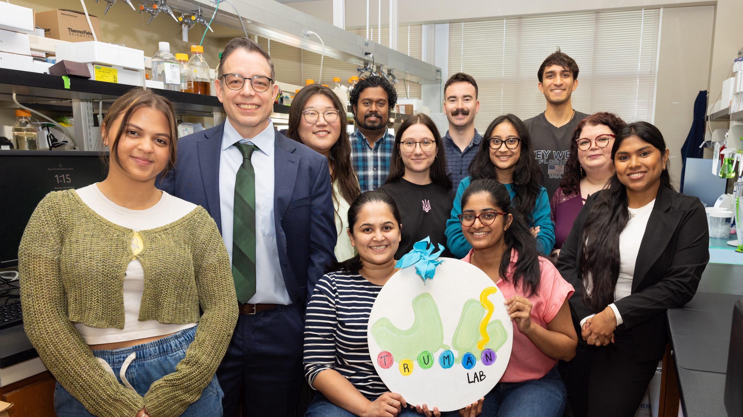 The Truman Lab members all posing for a photo in the biology lab. The group is standing in two rows, with two researchers sitting on stools in the front and holding a circular white sign with a blue bow on top. The sign has a white background and says 'Truman Lab' in colorful paint. All members are smiling at the camera.