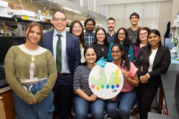 The Truman Lab members all posing for a photo in the biology lab. The group is standing in two rows, with two researchers sitting on stools in the front and holding a circular white sign with a blue bow on top. The sign has a white background and says 'Truman Lab' in colorful paint. All members are smiling at the camera.