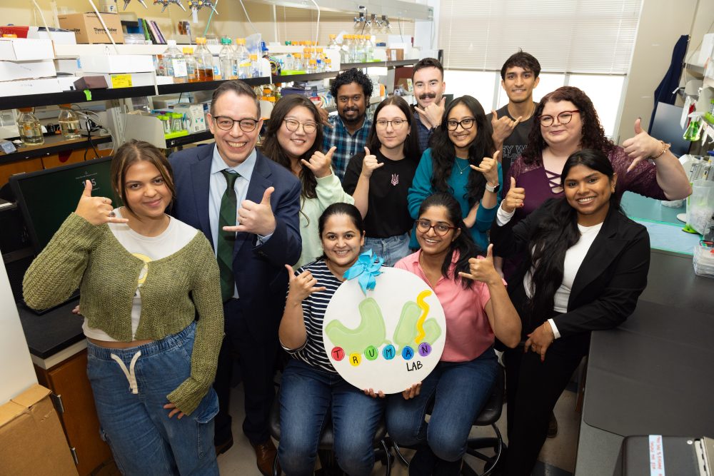 The Truman Lab members all posing for a fun photo in their lab. They are all smiling and holding the  "Go Niners" pickaxe hand gesture. 