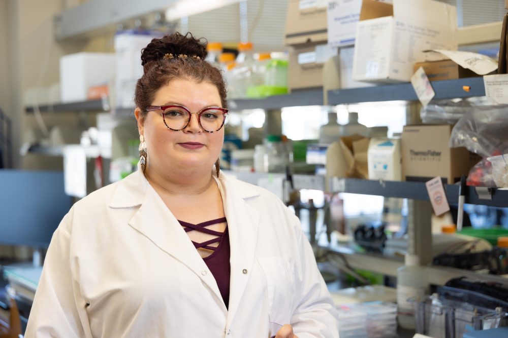 Mitchem posing in the lab looking directly at the camera. She is wearing a white lab coat, a maroon shirt, and maroon glasses.