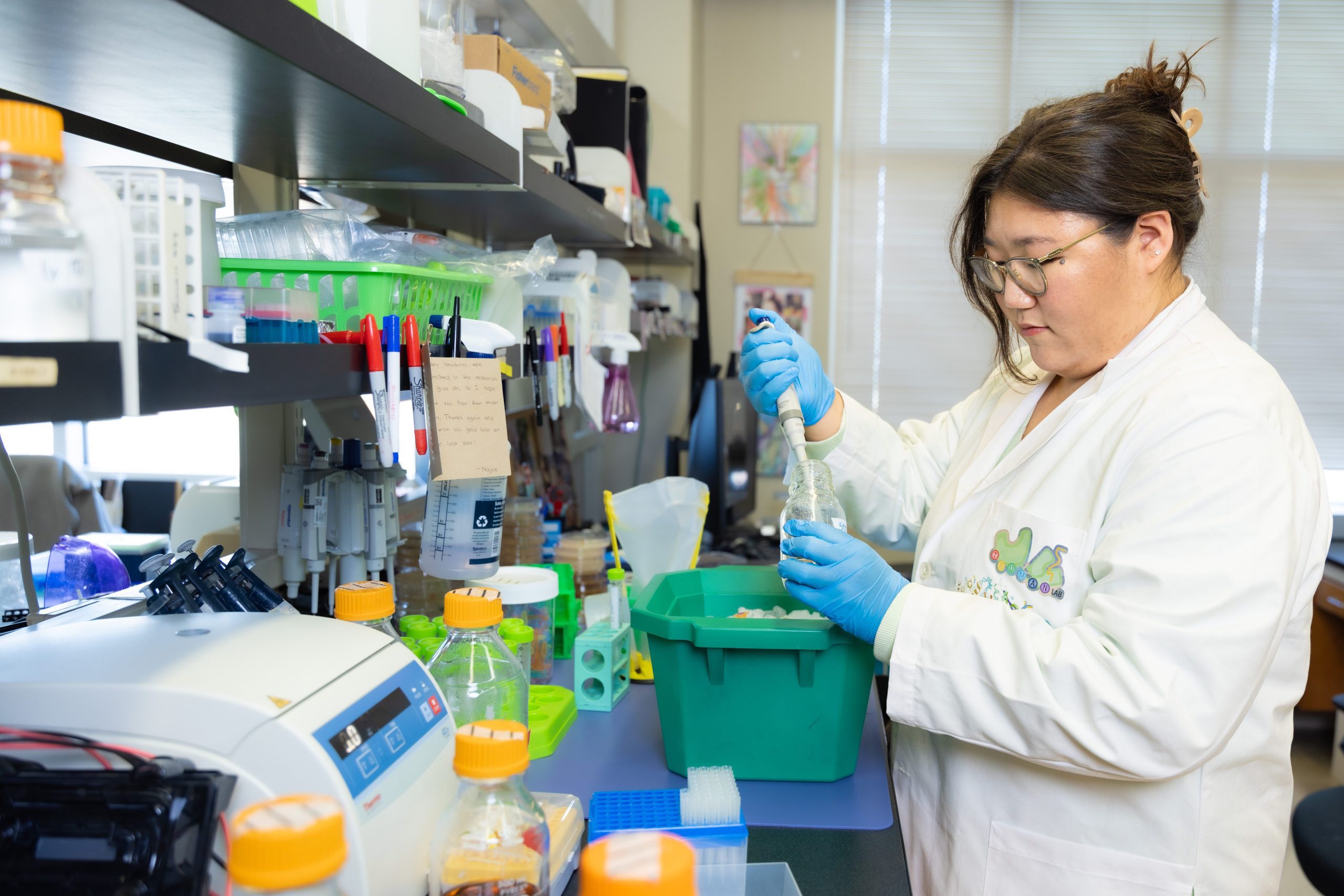 Choi working in a biology lab and wearing a white lab coat, blue protective gloves, and glasses. She is standing in front of a work counter covered with bottles and tools.