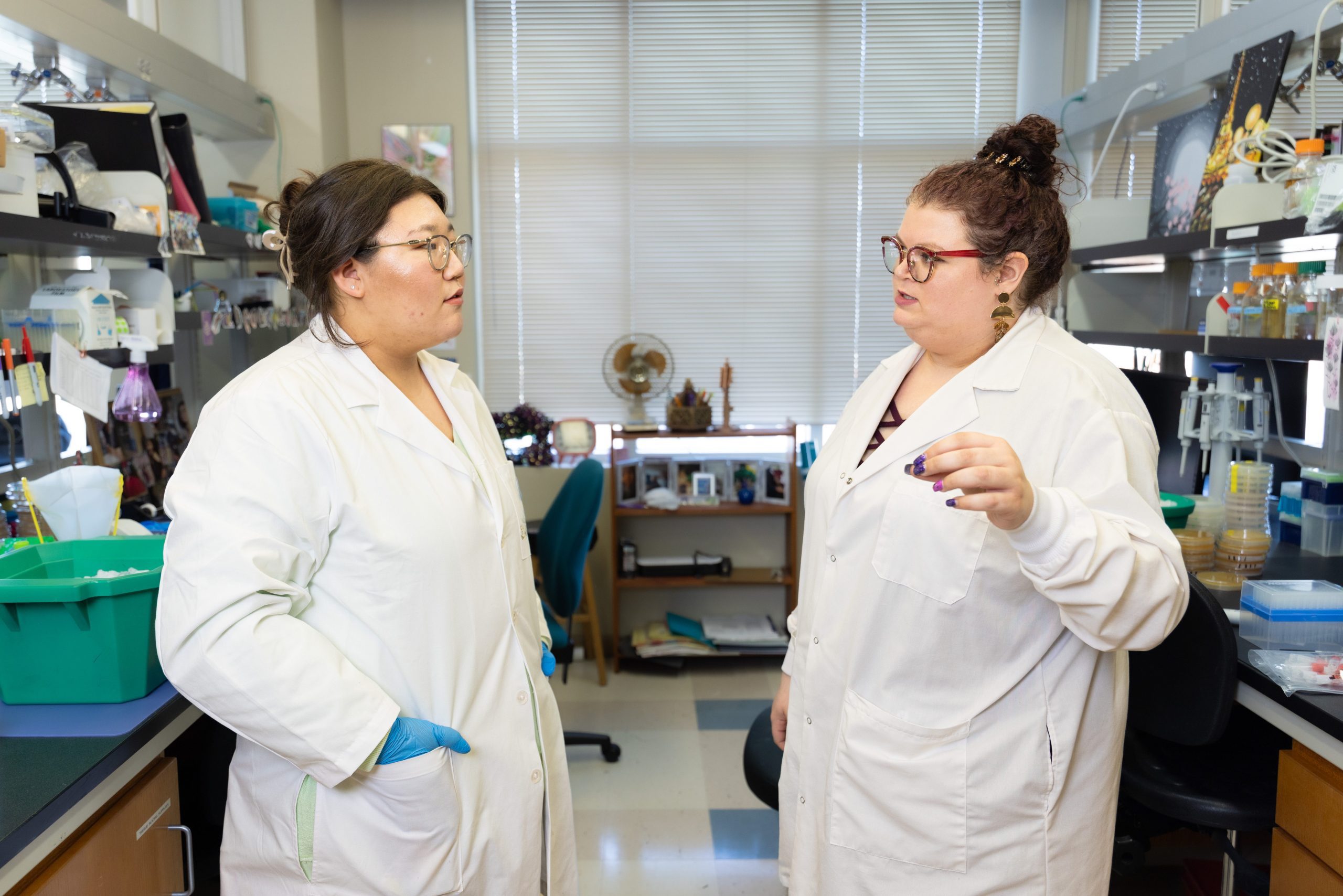 Choi and Mitchem standing in the aisle of the biology lab. Both are wearing white coats and appear to be having a serious discussion.