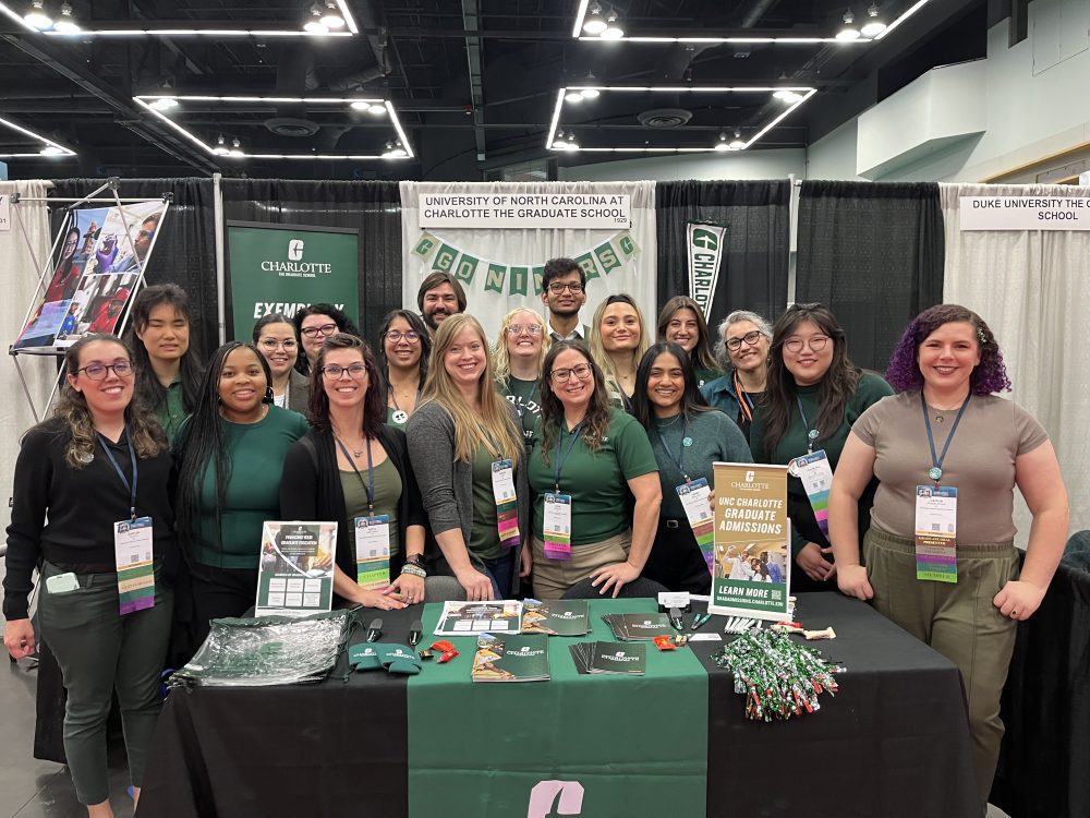 Eighteen members of Charlotte's chapter of SACNAS stand behind a table covered in black and green UNC Charlotte gear.