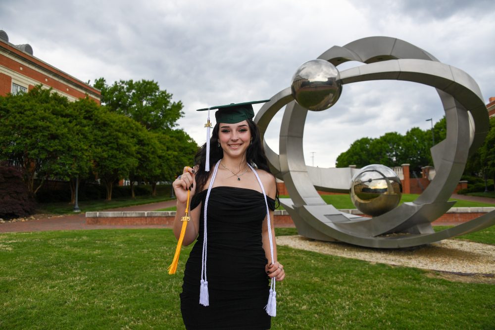 Allison Reed wears honor cords and her cap with one tassel while holding the other tassel for her second degree, standing in front of the Orbis sculpture.