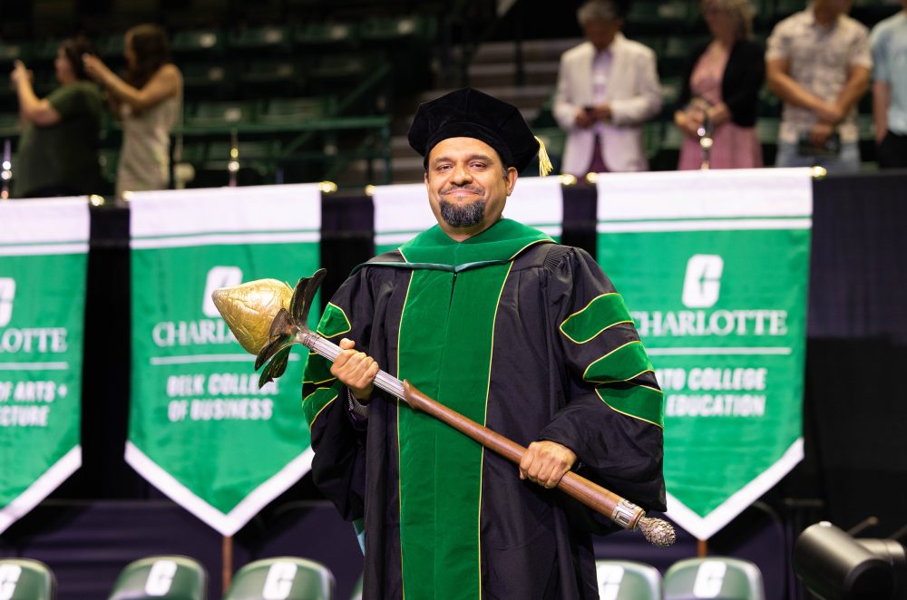 Juan Vivero-Escoto smiles on stage at commencement while holding the mace and wearing black and green graduation robes.
