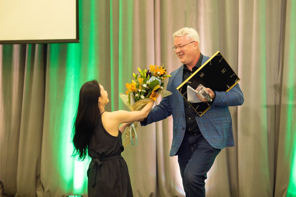 Kirill Afonin holds a gold framed award and is handed a large bouquet of flowers.