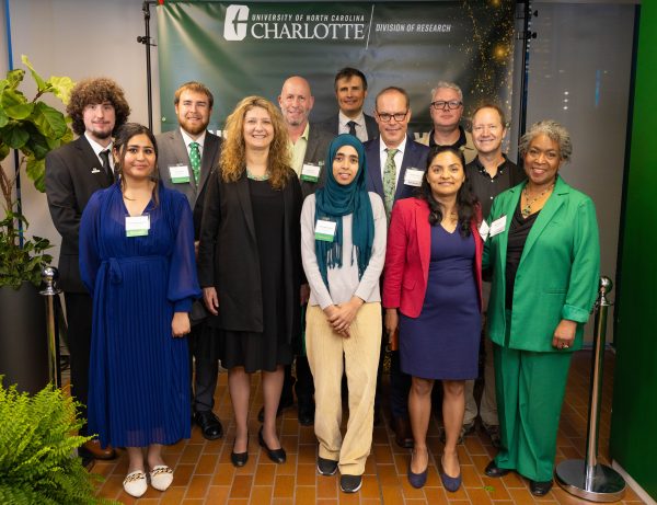 A group of smiling faculty and students from the Klein College of Science stand in front of a UNC Charlotte backdrop.