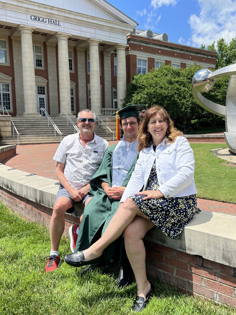 Ean Bass in his graduation cap and gown, sitting with his parents outside of Grigg Hall.