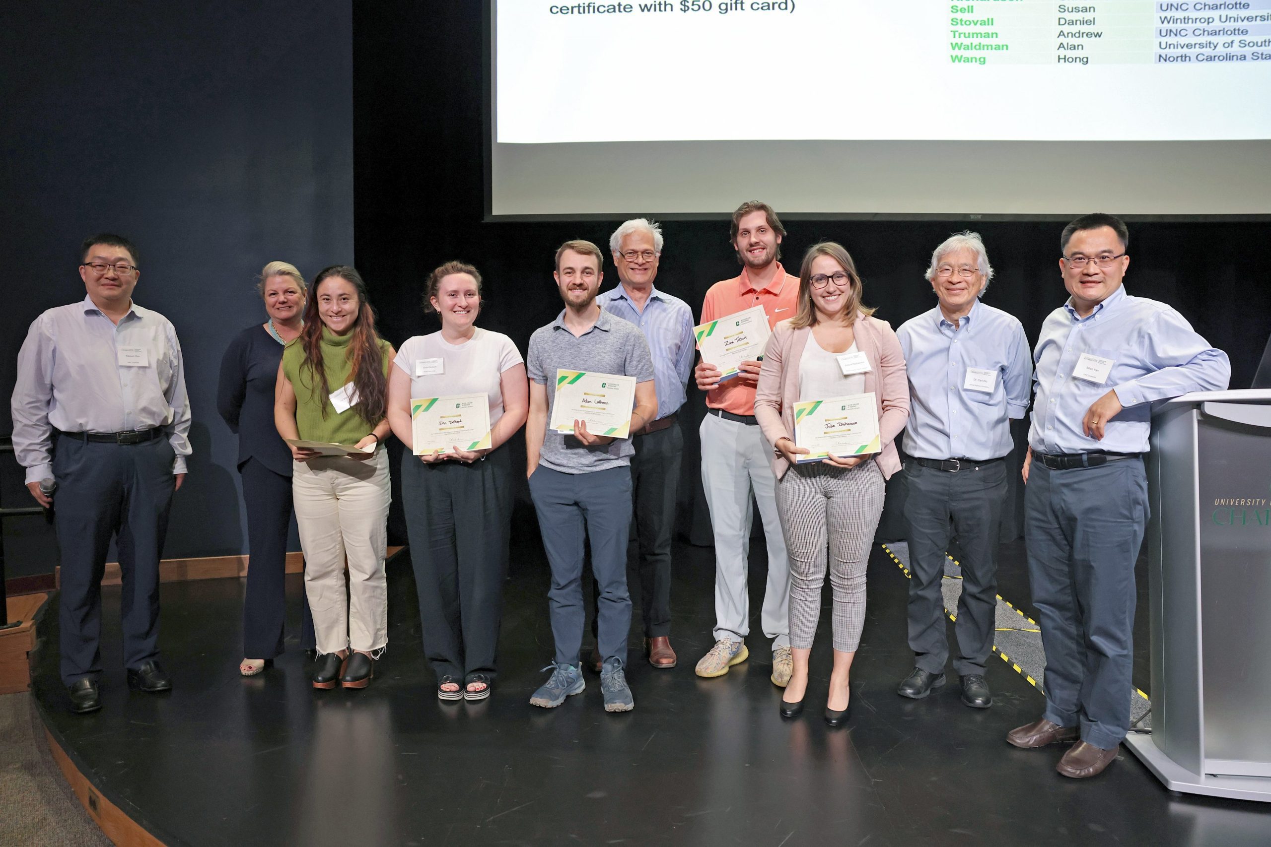 A group of researchers and symposium attendees on stage, with poster winners holding their certificates and smiling.