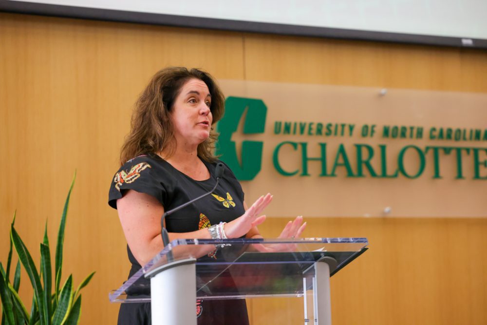 Tracy Dobson standing at a podium mid-presentation. Background includes green UNC Charlotte logo.