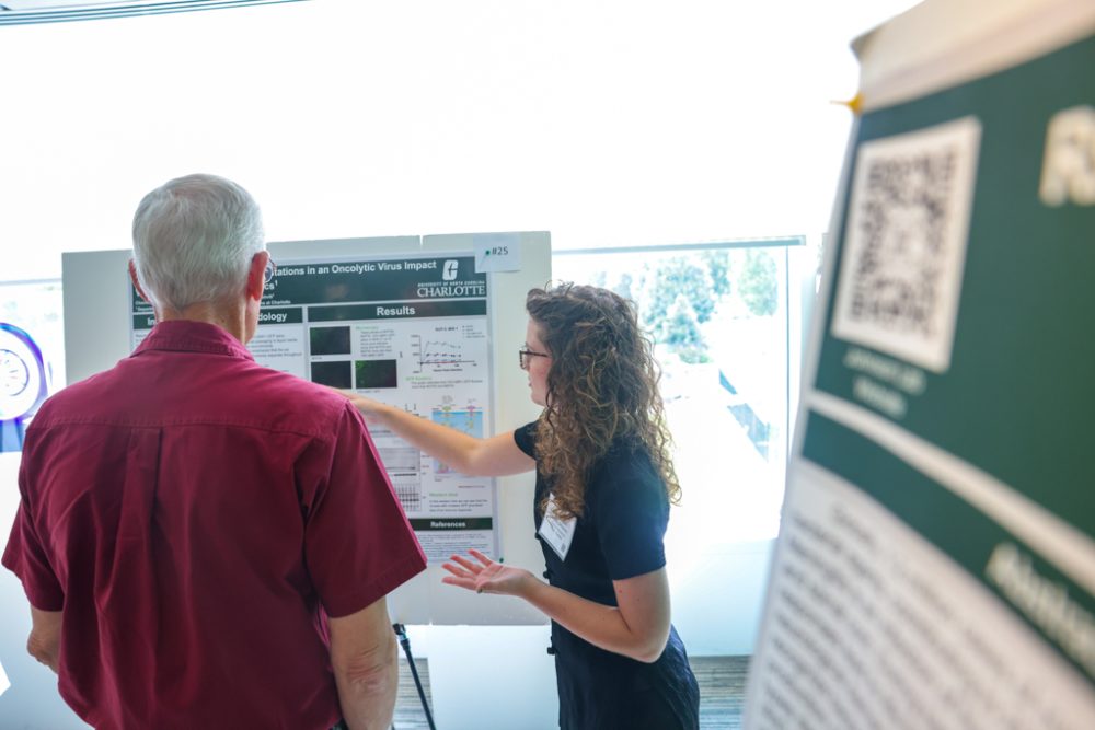 A student pointing at her research poster while an attendee watches. Another poster can be seen in the right foreground.