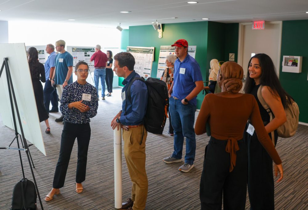 A student presenting to several event attendees. Additional posters and attendees can be seen in the background.