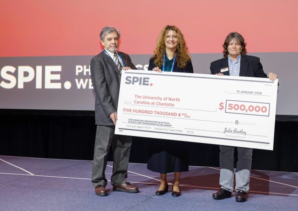 Three people hold a large ceremonial check made out to the University of North Carolina at Charlotte at the SPIE conference.