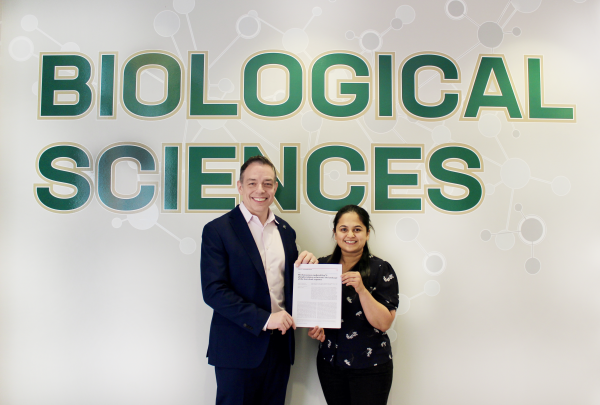Andy Truman and Siddhi Omkar smile in front of a "Biological Sciences" wall sign, holding a printed copy of their new article.