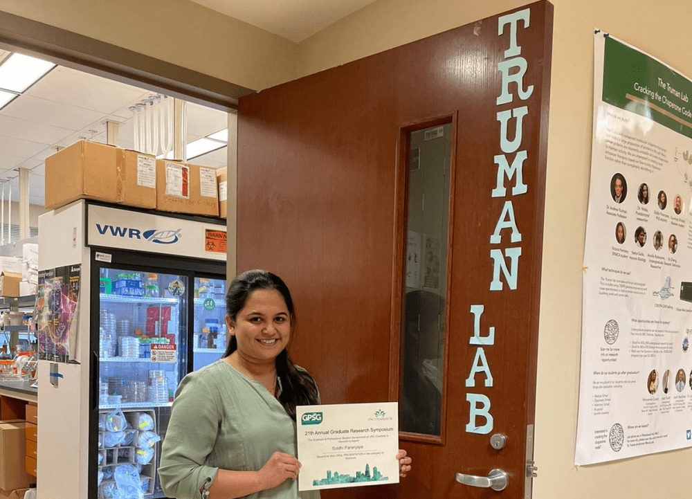 Omkar posing outside the lab door which reads "Truman Lab." She is holding a certificate from the 21st Annual Graduate Research Symposium.