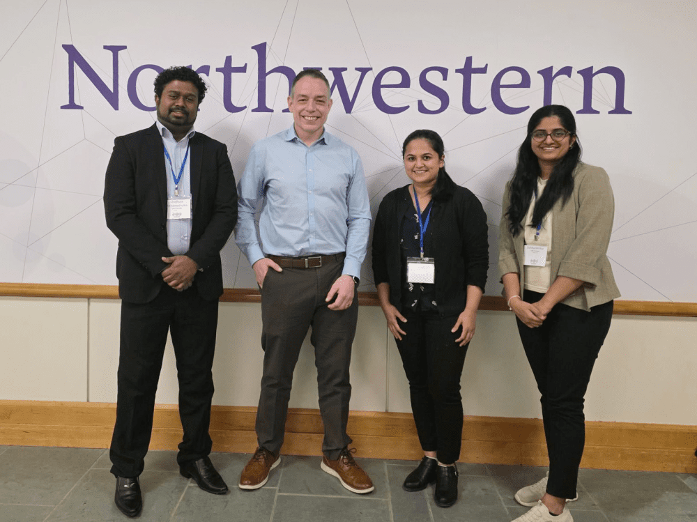Omkar, Truman, and two other members of the Truman Lab posing in front of a wall with a purple Northwestern University logo on it. 