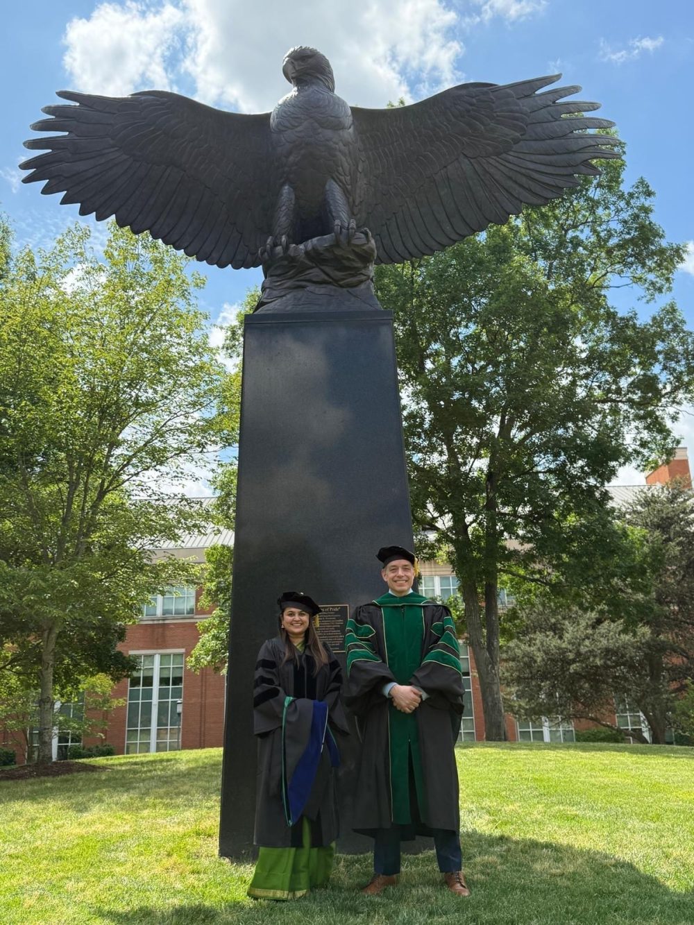 Omkar and Truman smile in front of the eagle statue behind Woodward Hall, wearing their Ph.D. caps and gowns.