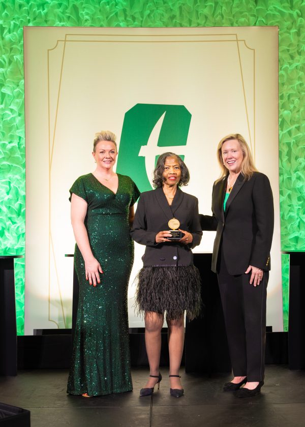 Robyn Massey holds her award on the UNC Charlotte stage, smiling and standing between Summer Baruth and Chancellor Sharon Gaber.