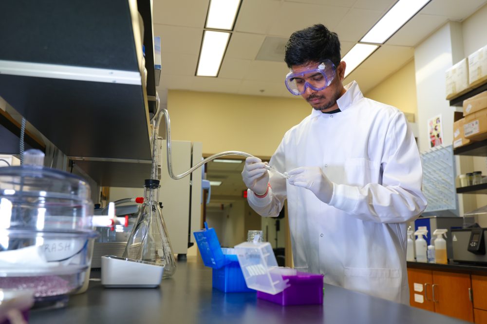 A researcher wears a lab coat, gloves and safety goggles, holding a tube attached to a flask.