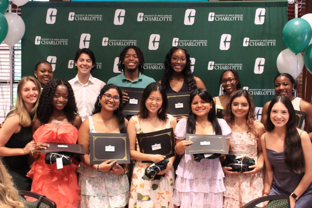 Ntasin and twelve Niner guides pose in front of UNC Charlotte backdrop holding certificates.