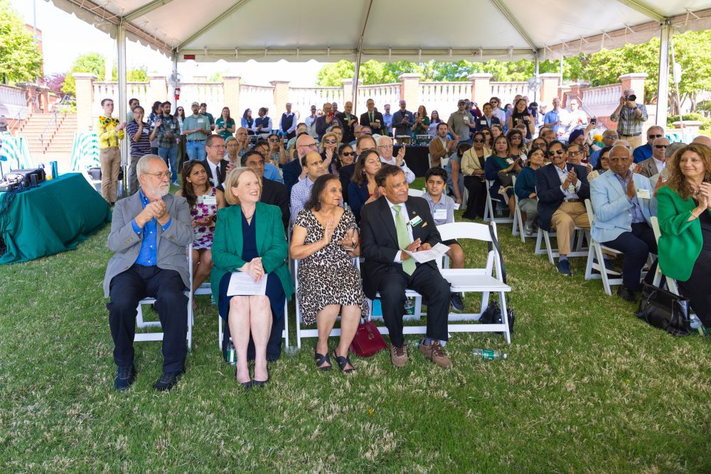 A large crowd is gathered under a tent on UNC Charlotte's campus, smiling and clapping. Dignitaries sit in the front row. Every chair is full and there is a crowd standing at the back.