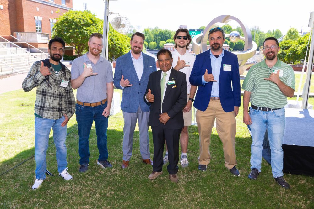 7 people stand smiling and showing pickaxe hand signs by the statue at Grigg Hall.