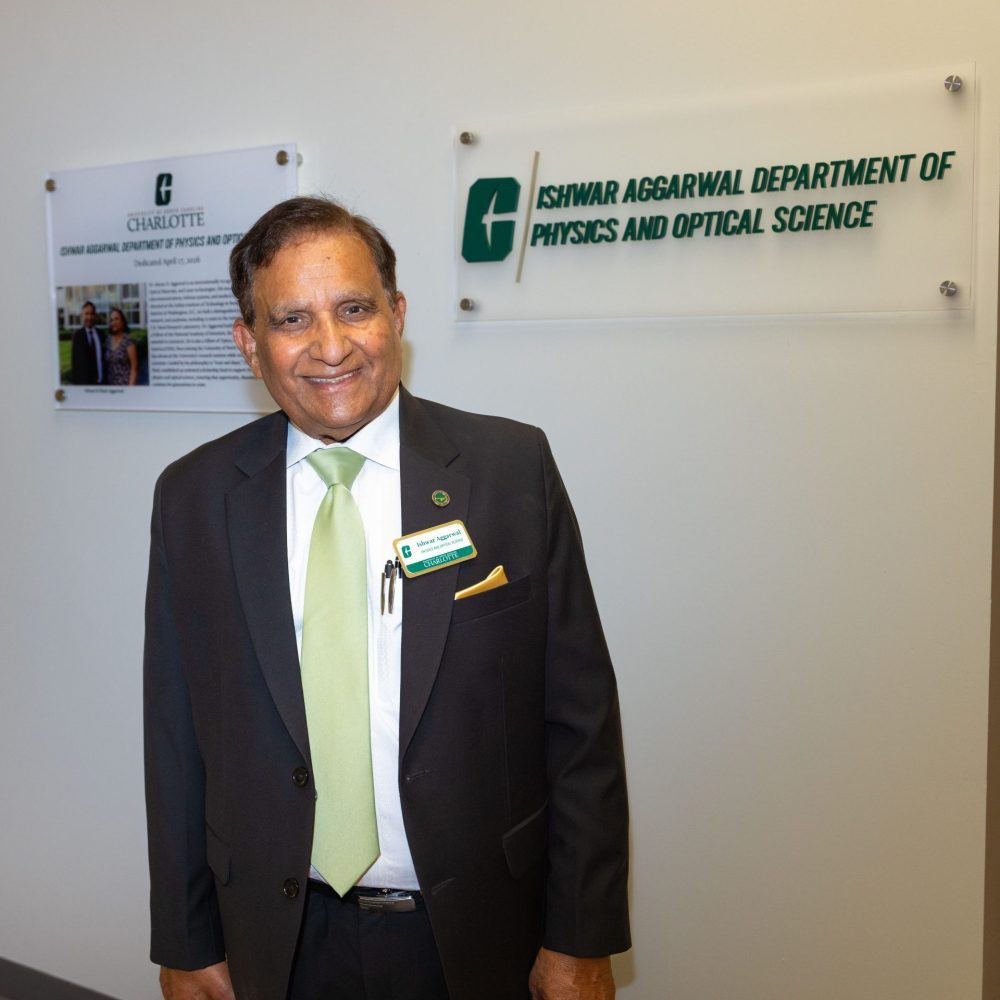 Ish Aggarwal wears a suit with a green tie and stands inside UNC Charlotte's Grigg Hall, in front of two new signs designating the Aggarwal Department of Physics and Optical Science.