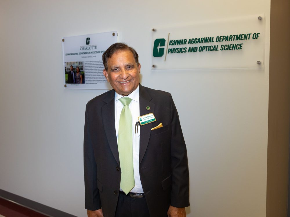 Ish Aggarwal wears a suit with a green tie and stands inside UNC Charlotte's Grigg Hall, in front of two new signs designating the Aggarwal Department of Physics and Optical Science.