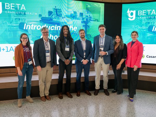 7 people, including the chosen healthcare innovators, stand in front of screens with the gBeta logo.