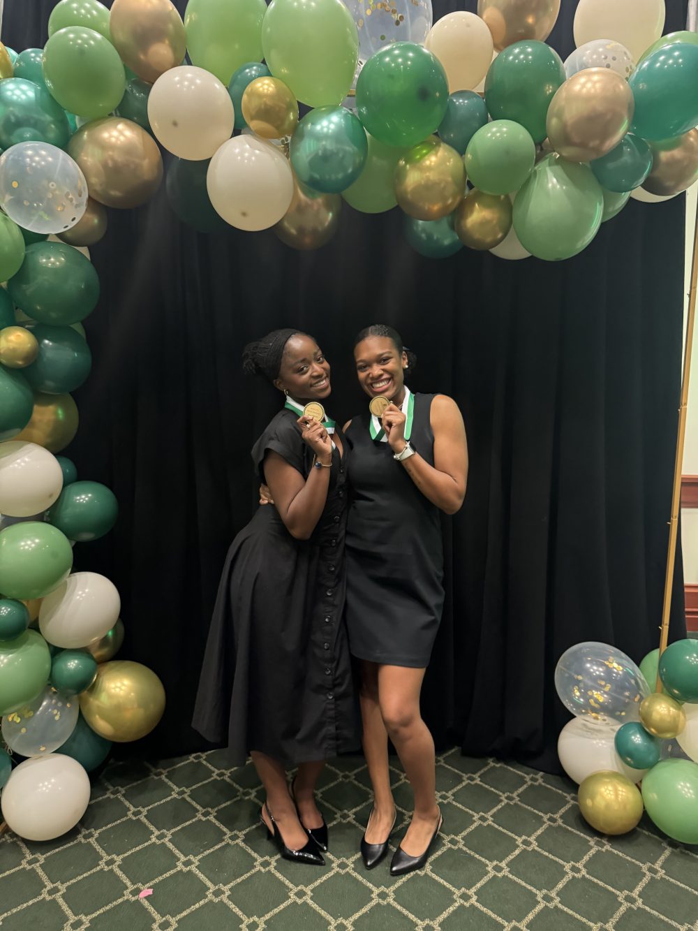 Ntasin and friend pose with their medals in front of a black cloth backdrop and a green and gold balloon arch. 