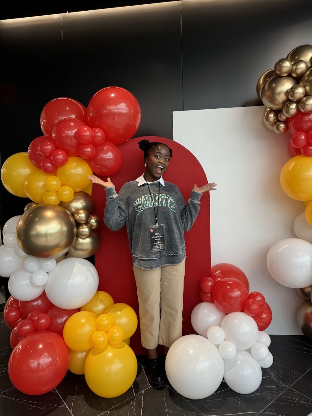 Ntasin wearing an UNC Charlotte sweatshirt in front of a red, yellow, gold and while balloon formation.