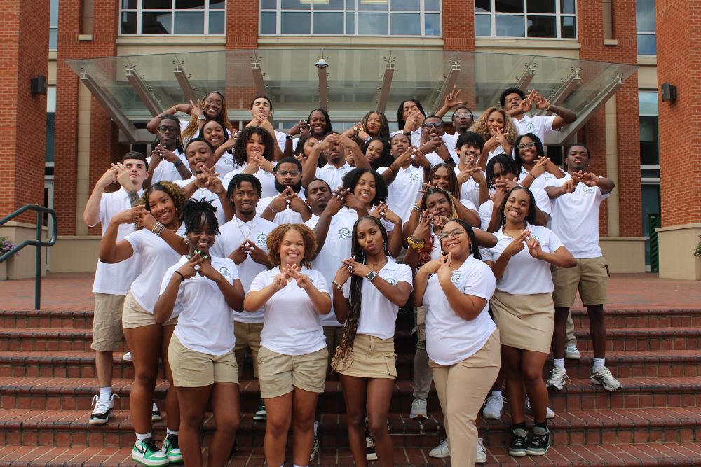 A group of students in white t-shirts and khakis pose in front of a UNC Charlotte building.
