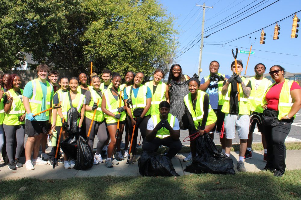 A large group wearing high-vis vests and carrying trash bags and grabbing equipment pose for a photo on a sidewalk in Charlotte.
