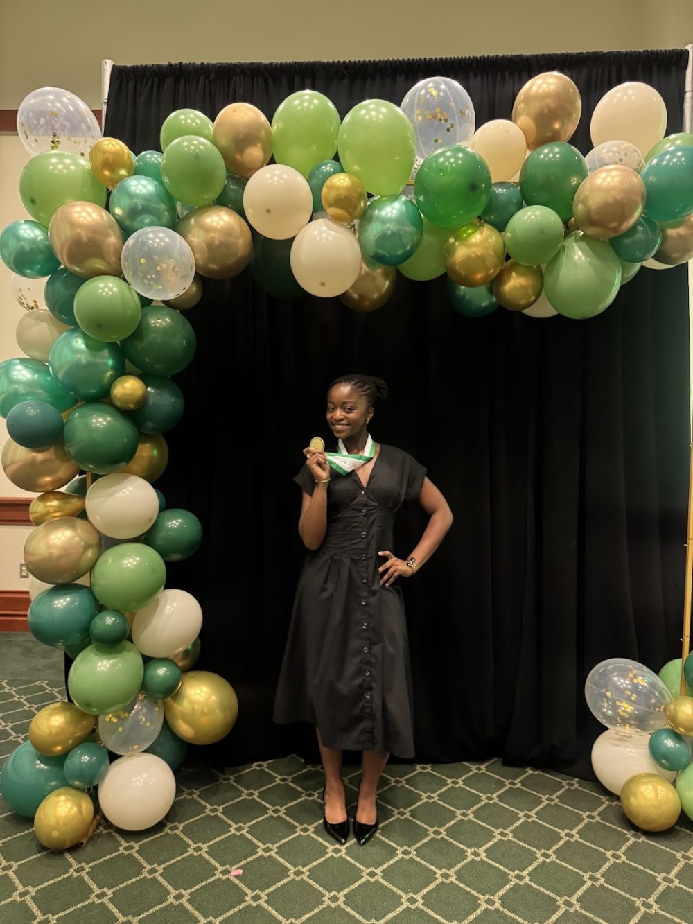 Ntasin holds her medal in front of a black cloth backdrop and a green and gold balloon arch.