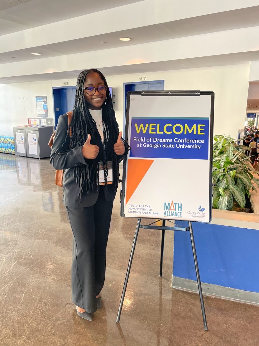 Ntasin giving a thumbs up to the camera alongside a sign that says "Welcome Field of Dreams Conference at Georgia Stat University."