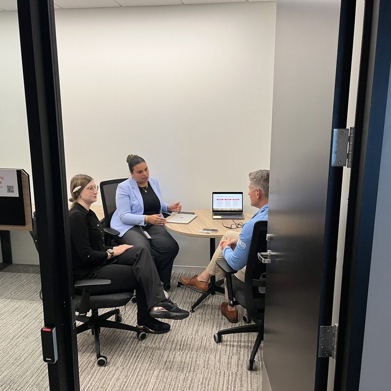 Three people sit inside a room chatting around a table, as seen through a doorway.