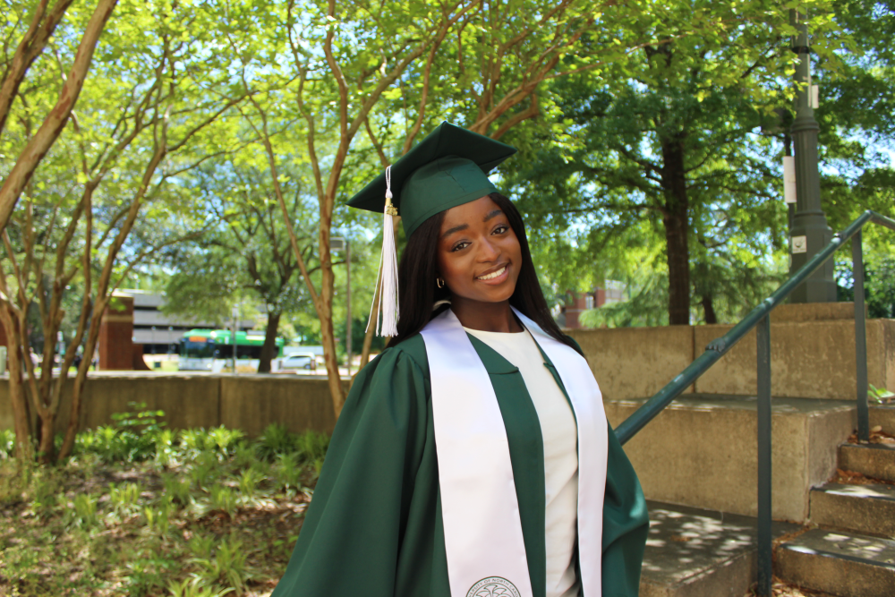 Ntasin posing in her cap and gown outside of the Fretwell building next to a staircase. A few trees and a bus can be seen in the background.