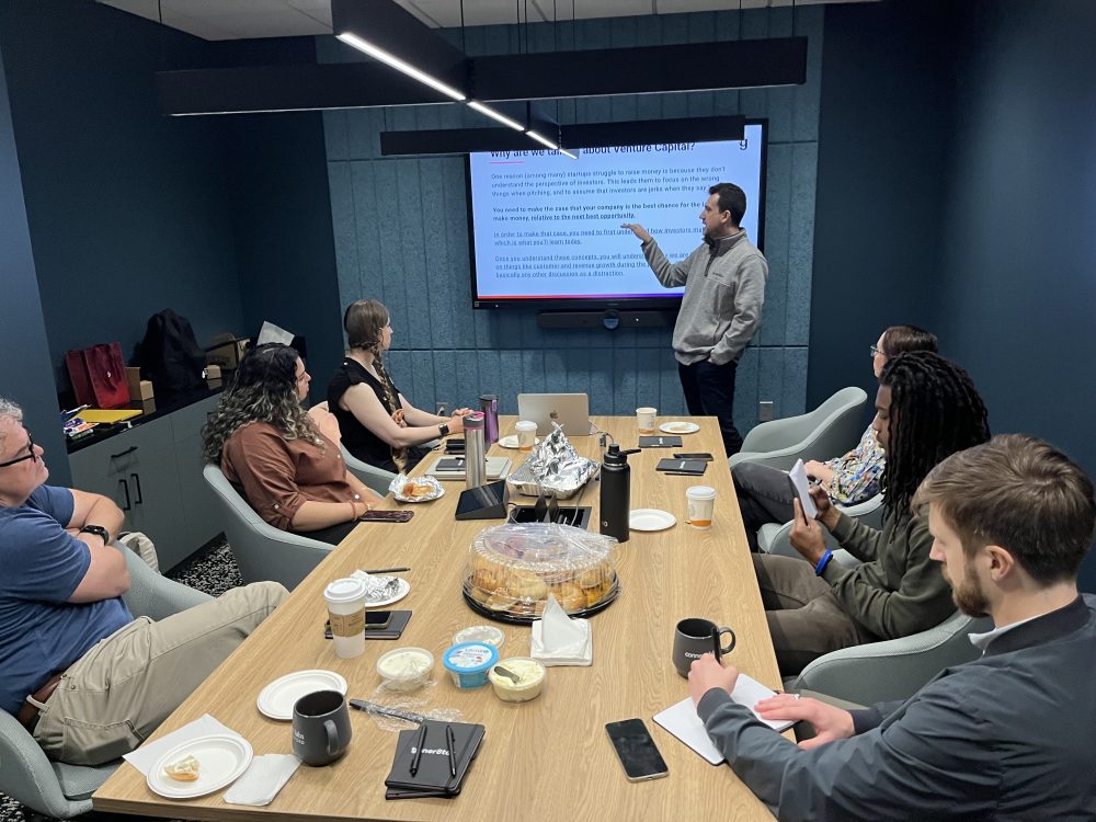 A group of people sit at a conference table, looking at the presenter at the front of the room pointing to the screen.