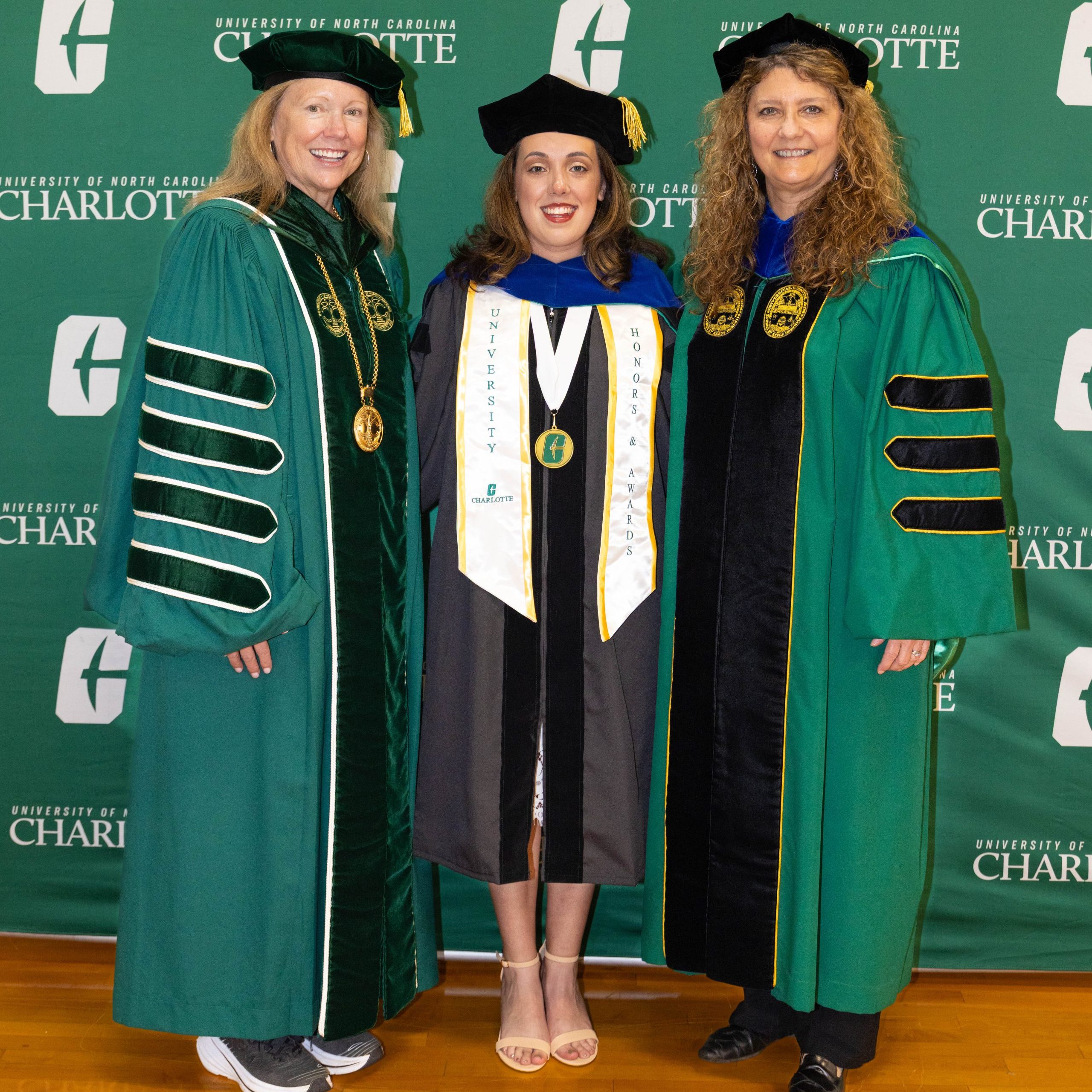 3 people in graduation regalia stand in front of a UNC Charlotte backdrop. UNC Charlotte Chancellor Sharon L. Gaber, Kaitlin Klotz, Klein College of Science Founding Dean Bernadette Donovan-Merkert.