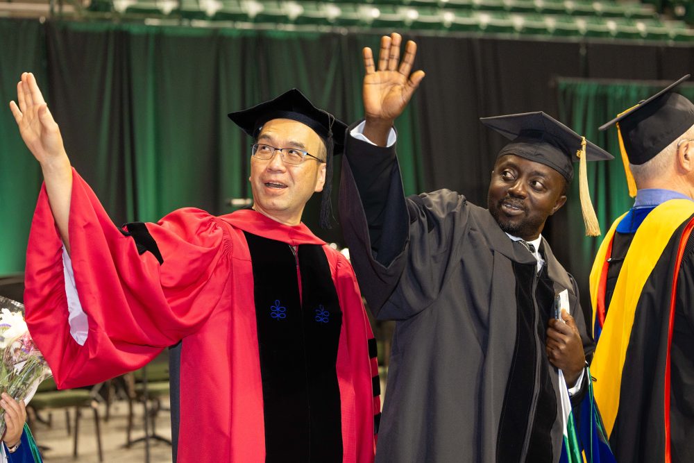 A Ph.D. student and their advisor wearing graduation regalia wave to the crowd.