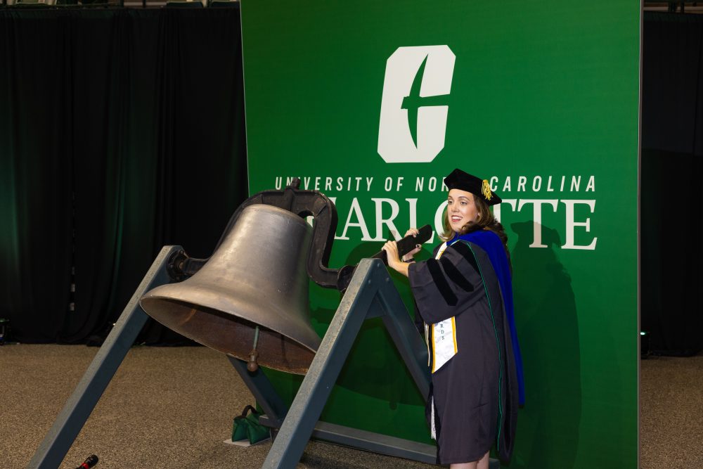 Kaitlin Klotz in doctoral graduation regalia rings the large old bell in front of a UNC Charlotte backdrop.