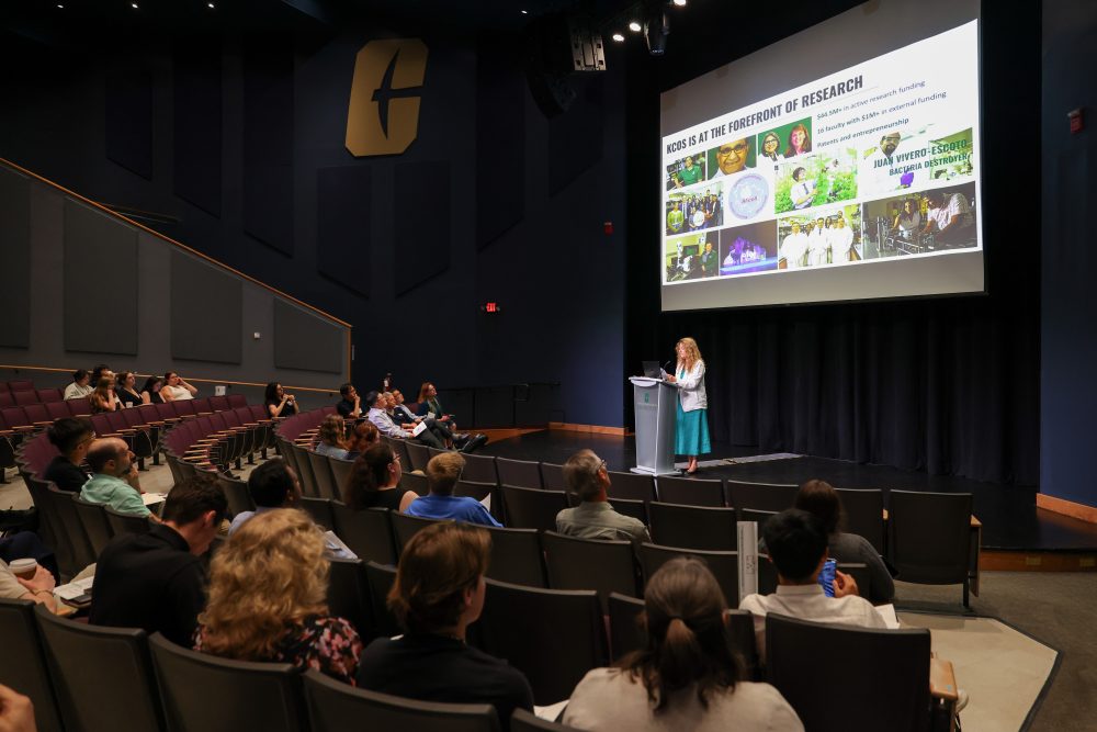 Bernadette Donovan-Merkert, founding dean of the Klein College of Science, speaking on stage with a presentation on a screen above her. The back of the audience can be seen in the foreground of the image.