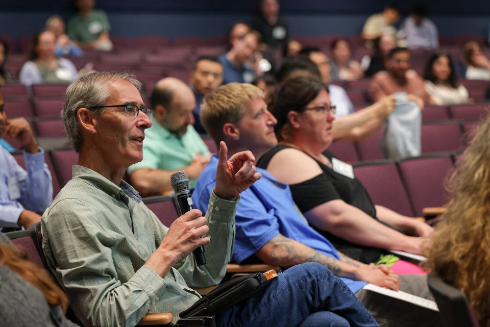 An audience member asks a question while holding a microphone, while others listen and look ahead.
