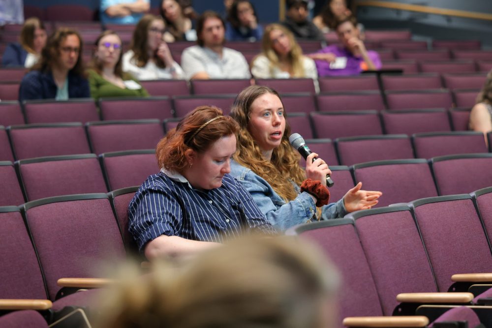 Two audience members. One is speaking into a microphone and asking a question, while the other writes notes.