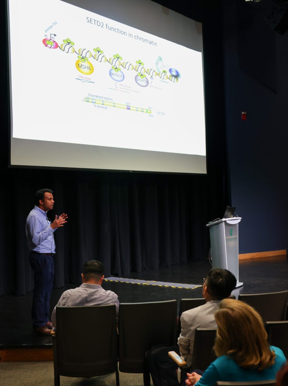 Khan on stage presenting his PowerPoint presentation. The back of audience members can be seen in the foreground.